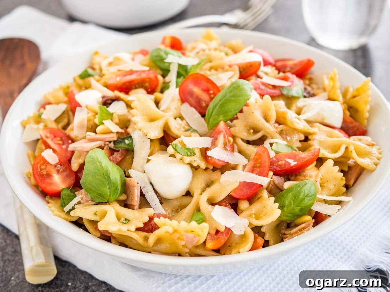 Zesty Italian Pasta Salad 7 A white bowl with pasta salad with italian dressing, with tomato, mozzarella, basil leaves and shavings of Parmiggiano. The bowl is sitting on a white dishtowel with forks, a glass of water and salad tongs in the background.
