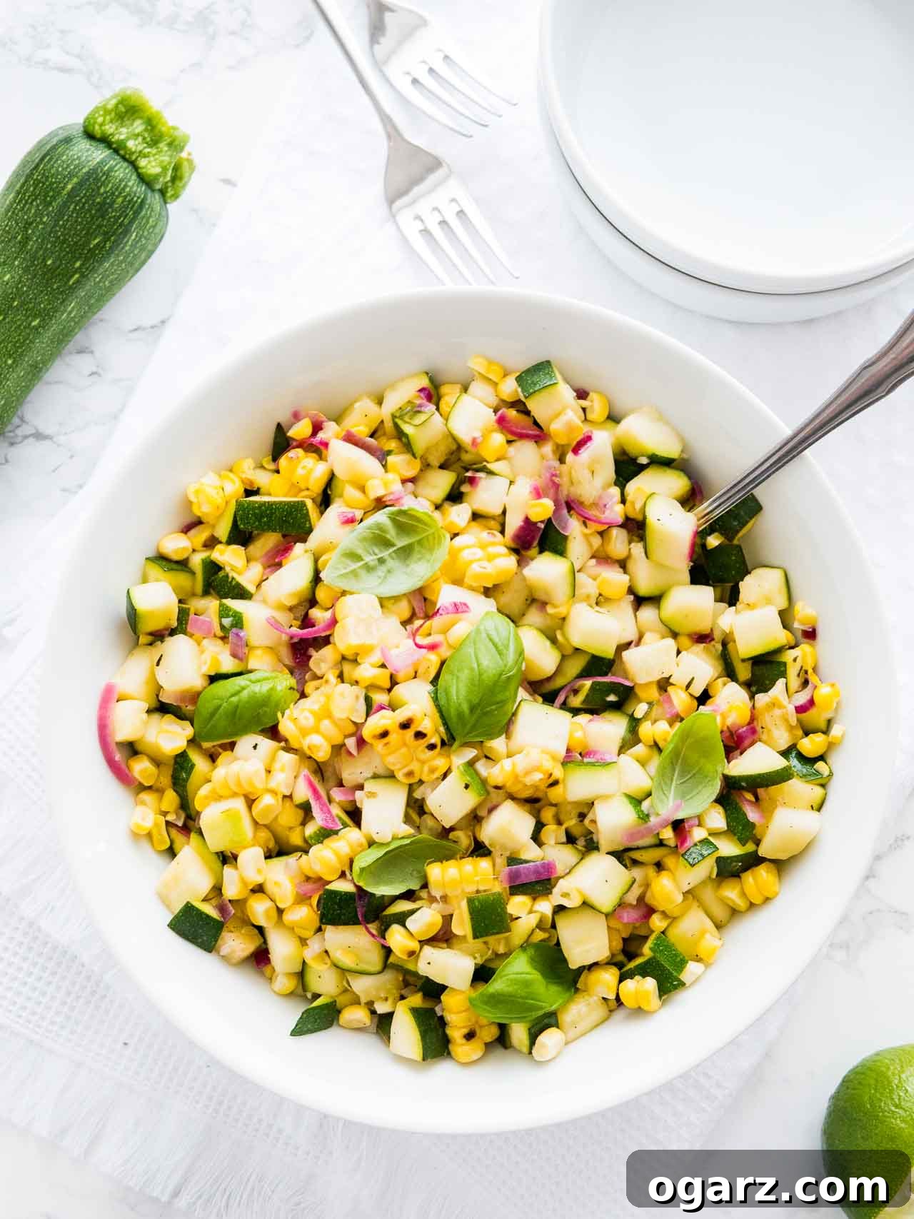 Zucchini Corn salad in a white bowl, garnished with basil, with a spoon in it on a white table with a fork and a zucchini next to it.