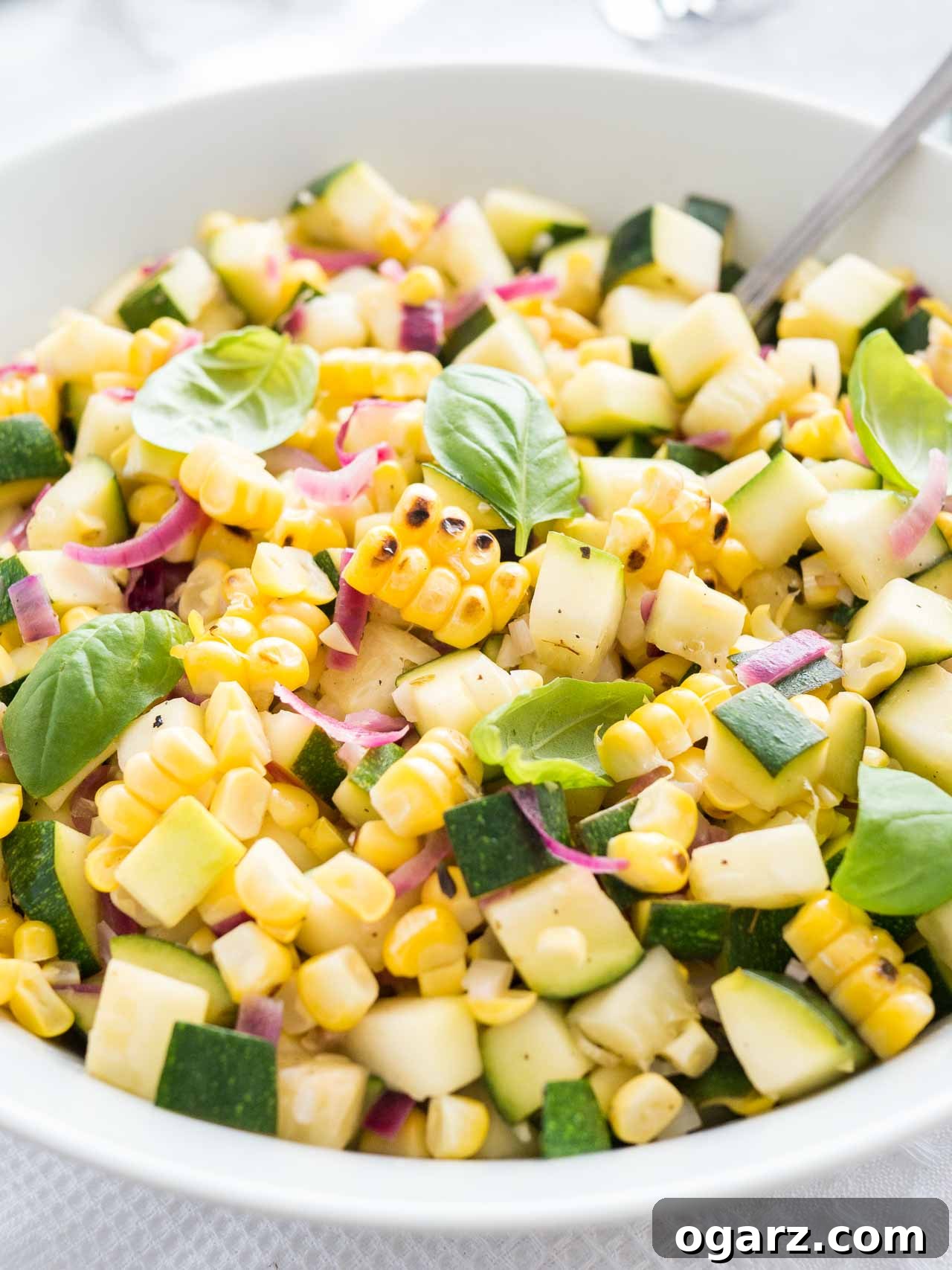 Close-up of roasted corn zucchini salad with onions in a white bowl, garnished with basil, with a spoon in it.