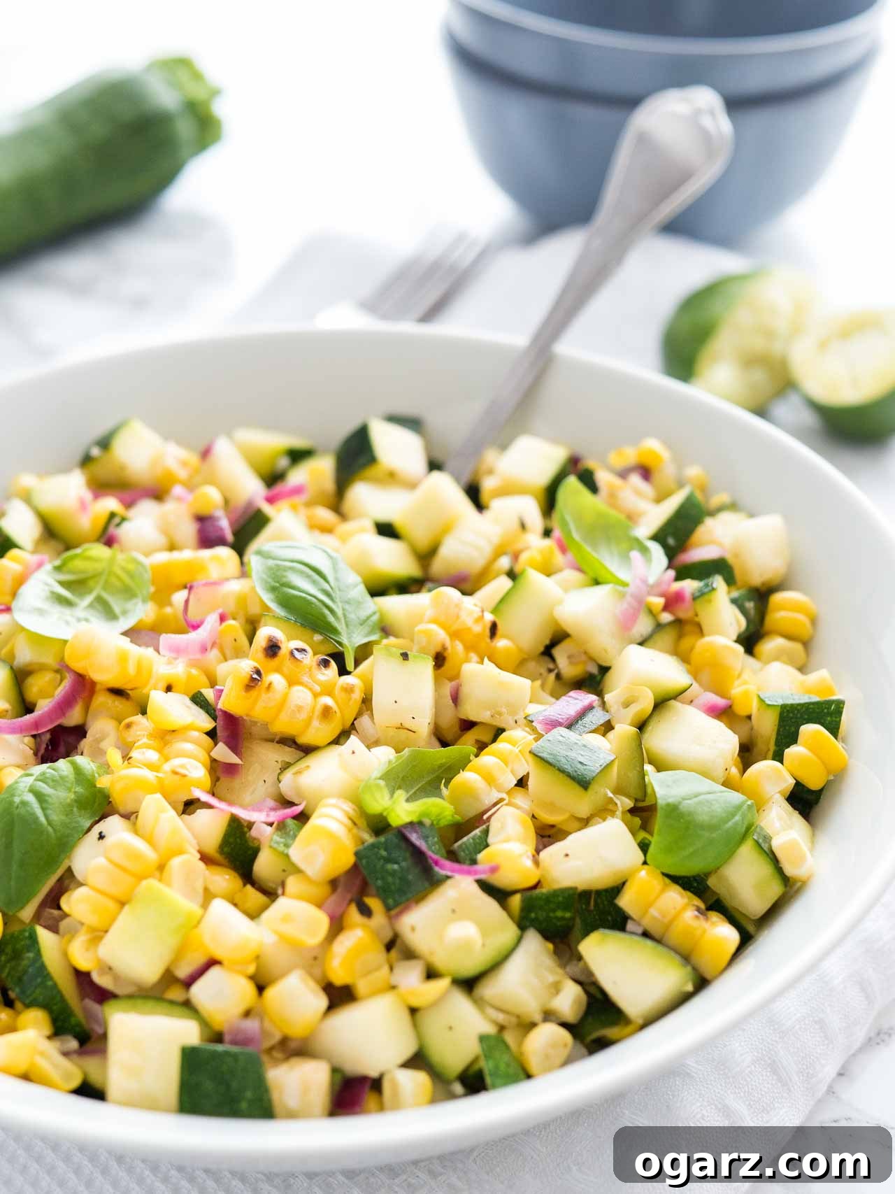 Roasted corn zucchini salad with onions in a white bowl, garnished with basil, with a spoon in it on a white table with a fork, zucchini and squeezed limes in the background.