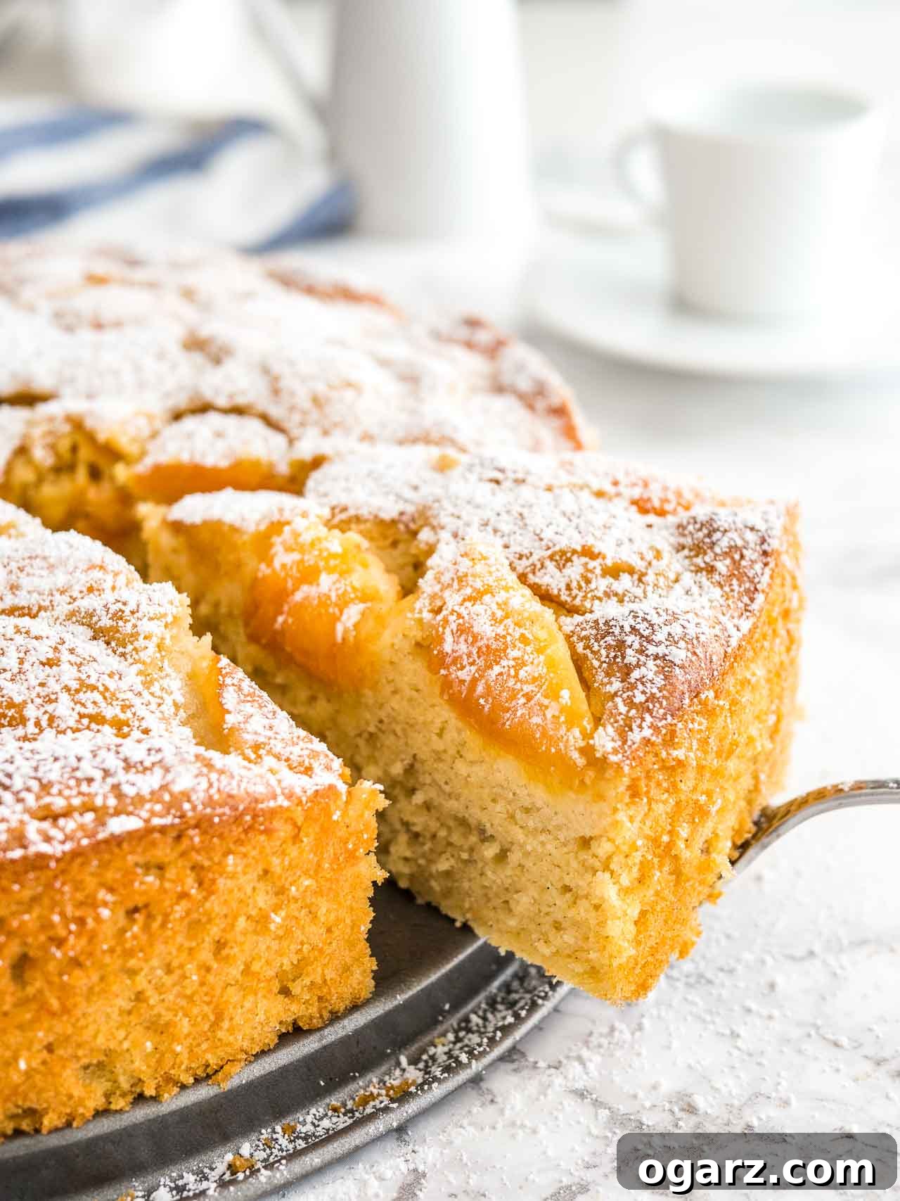 Golden Apricot Delight 2 A beautifully baked apricot cake on a springform bottom, with a slice being lifted out by a cake server. White coffee cups are visible in the soft-focus background.