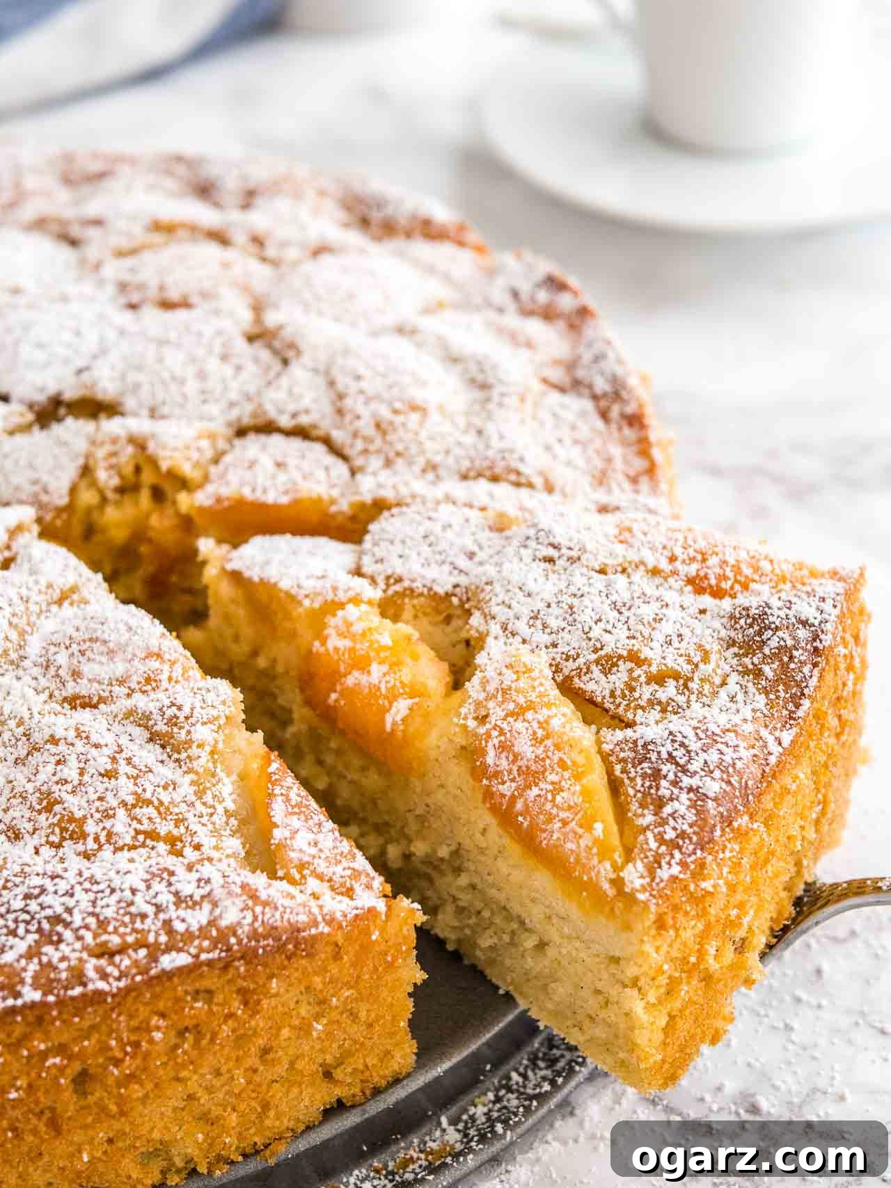 Golden Apricot Delight 5 Close-up of an apricot cake on a springform bottom, with a cake server lifting out a piece and white coffee cups in the background.