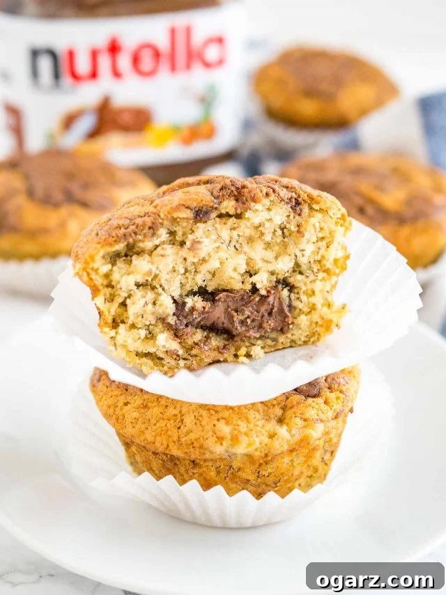 Close-up of two banana Nutella muffins stacked on a white plate, with more muffins and a Nutella jar in the soft-focus background. A large bite has been taken from the top muffin, revealing its moist interior and chocolate-haznut filling.