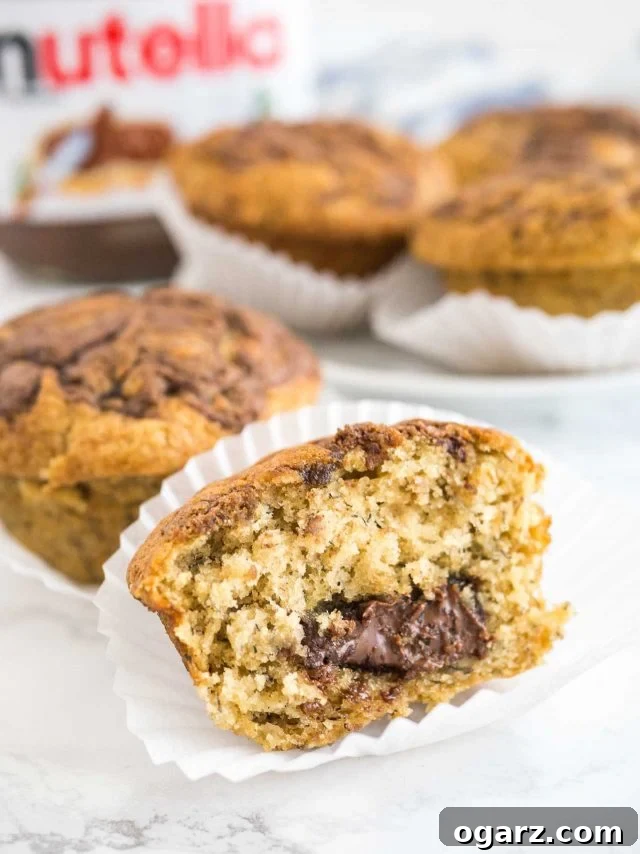 Close-up of a single banana Nutella muffin in its paper liner, with a large bite missing from its side, exposing the rich, dark Nutella core. More muffins and a jar of Nutella are visible in the blurred background.
