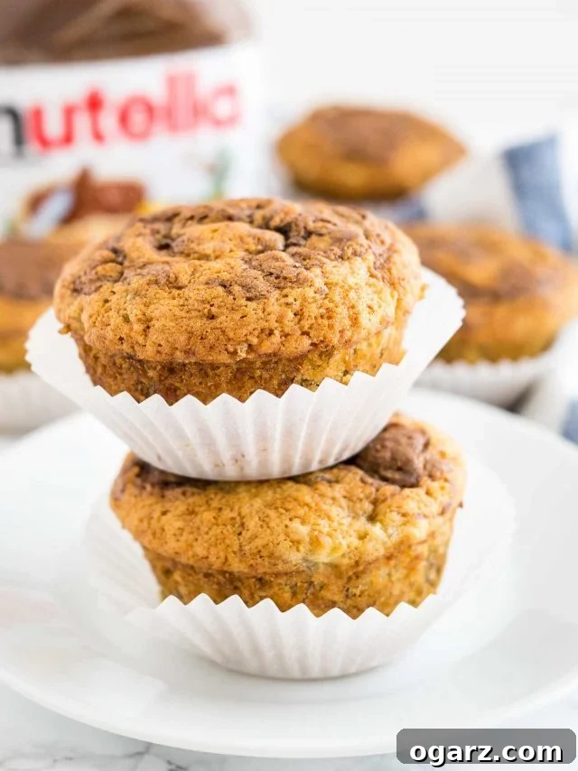 Close-up of two banana Nutella muffins stacked on top of each other on a pristine white plate. The muffins appear golden brown and perfectly domed, with visible Nutella swirls on top. More muffins and a Nutella jar are blurred in the background.