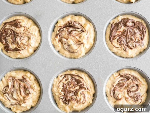 Top-down shot of a grey muffin pan, with 12 muffin cups filled with unbaked banana Nutella muffin batter. The Nutella swirls are visible on the surface of the batter, ready for baking.