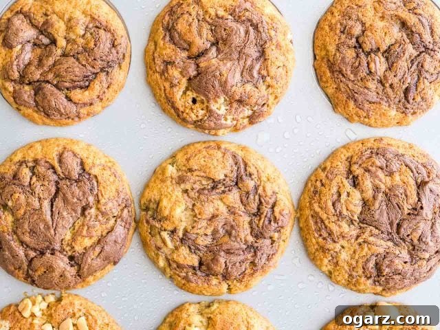 Top-down shot of a grey muffin pan, now filled with 12 golden-brown, freshly baked Banana Nutella Muffins, puffed high with perfect domed tops. The Nutella swirls are caramelized on the surface.