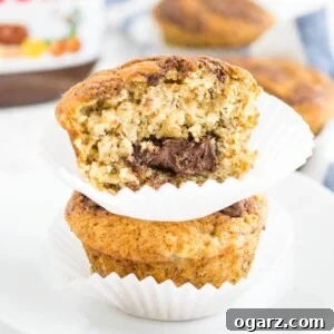 Two banana Nutella muffins stacked on top of each other on a white plate, with more muffins and a jar of Nutella in the background. The top muffin has had a big bite taken out of it.