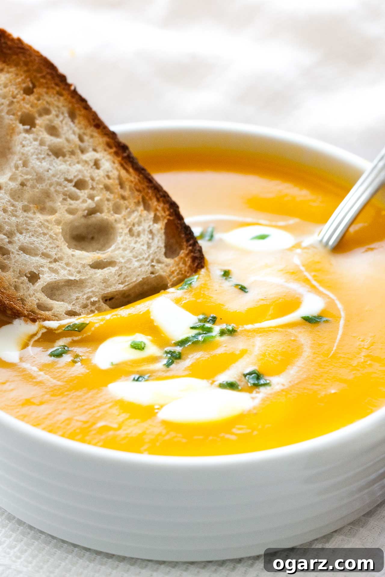 Silken Butternut Squash Delight 2 Close-up of a white bowl with butternut squash soup, with dollops of sour cream and chives, a spoon in it and a toasted slice of bread. It is sitting on a white dish towel next to some toasted slices of bread.