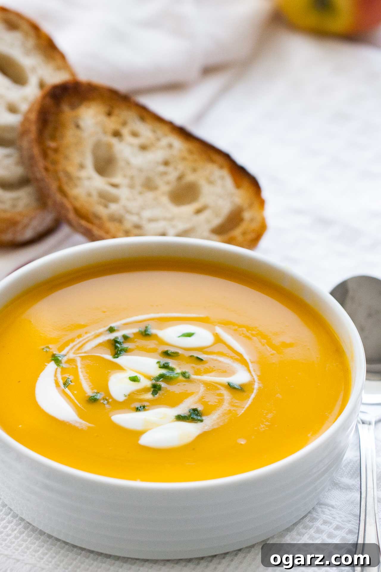 Silken Butternut Squash Delight 4 A white bowl with butternut squash soup, with dollops of sour cream and chives. It is sitting on a white dish towel and there's a spoon and toasted slices of bread next to it.