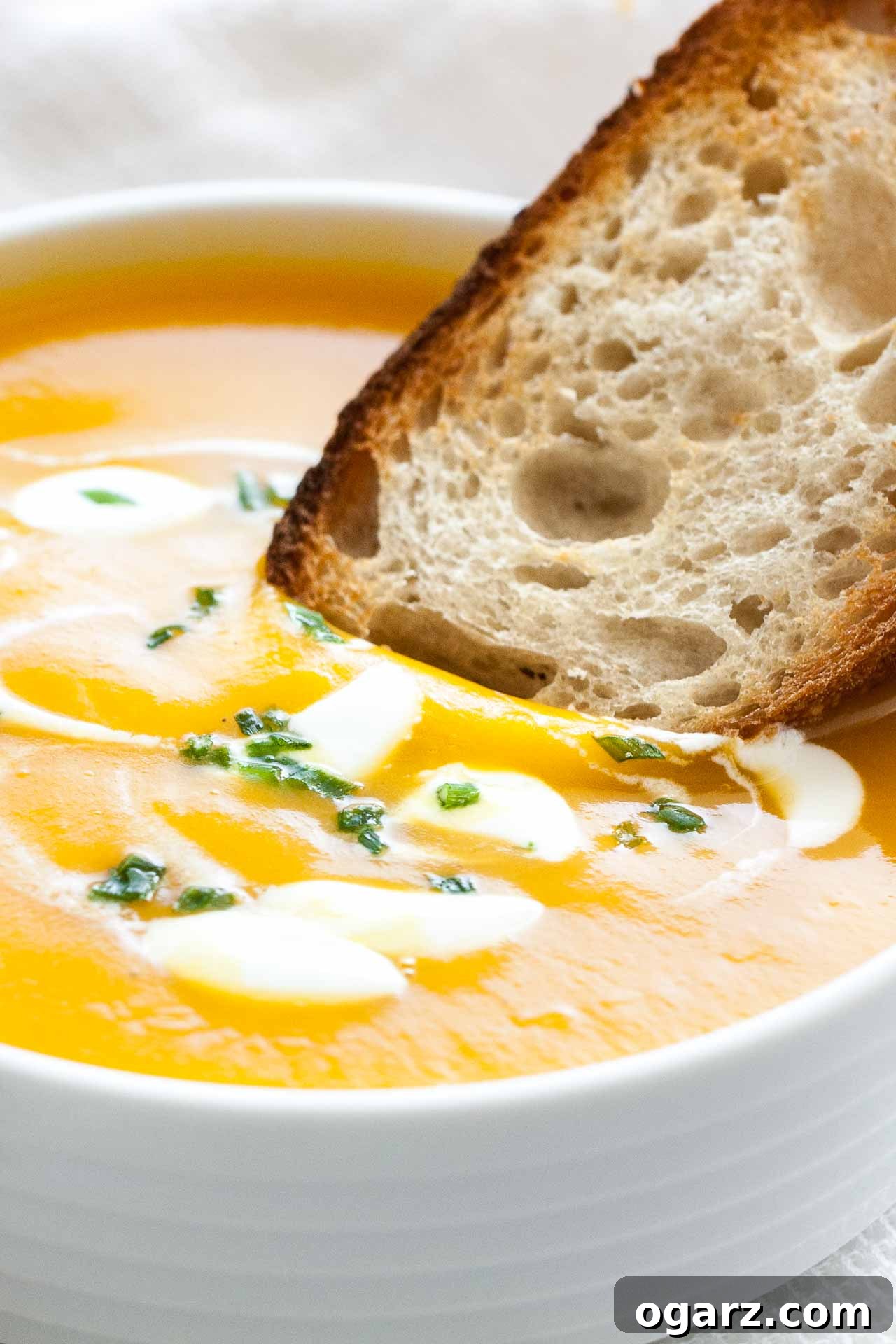 Silken Butternut Squash Delight 5 Close-up of a toasted slice of bread being dipped into butternut squash soup with dollops of sour cream and chives in a white bowl.