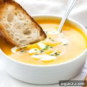 A white bowl with butternut squash soup, with dollops of sour cream and chives, a spoon in it and a toasted slice of bread. It is sitting on a white dish towel next to some toasted slices of bread.