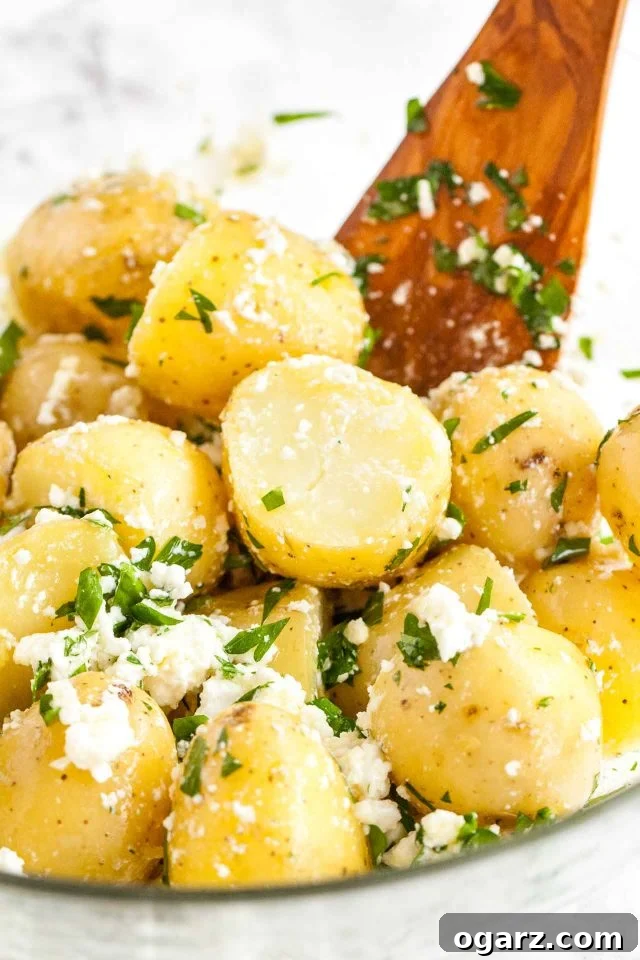 A close-up view of a glass serving bowl filled with Greek-Style Boiled Potatoes, generously topped with crumbled feta and vibrant green parsley, with a wooden spatula ready for serving.