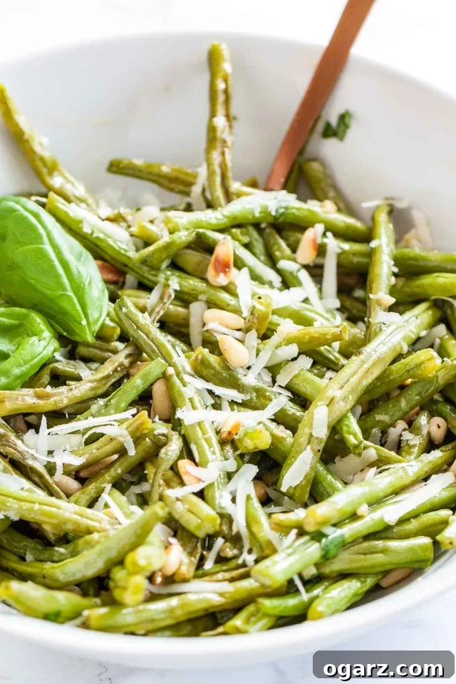 Close up of a white bowl of oven-roasted green beans topped with roasted pine nuts, Parmigiano and basil leaves, ready to serve.
