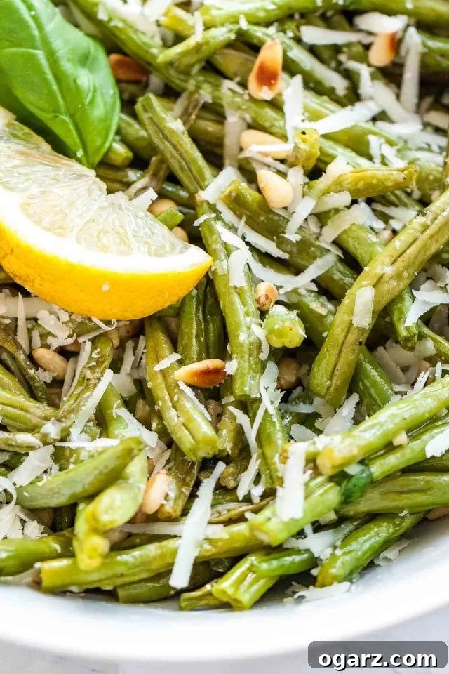 Close up of oven-roasted green beans topped with roasted pine nuts and Parmigiano, garnished with a basil leaf and a wedge of lemon, ready to be served on a platter.