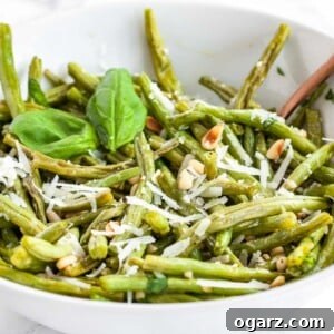 A white bowl of oven-roasted green beans topped with roasted pine nuts and Parmigiano, garnished with a basil leaves.