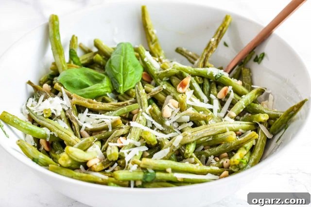 Serve up these delicious Parmesan Oven Roasted Green Beans for your next family gathering or holiday. A white bowl of oven-roasted green beans topped with roasted pine nuts and Parmigiano, garnished with a basil leaves, presented beautifully.