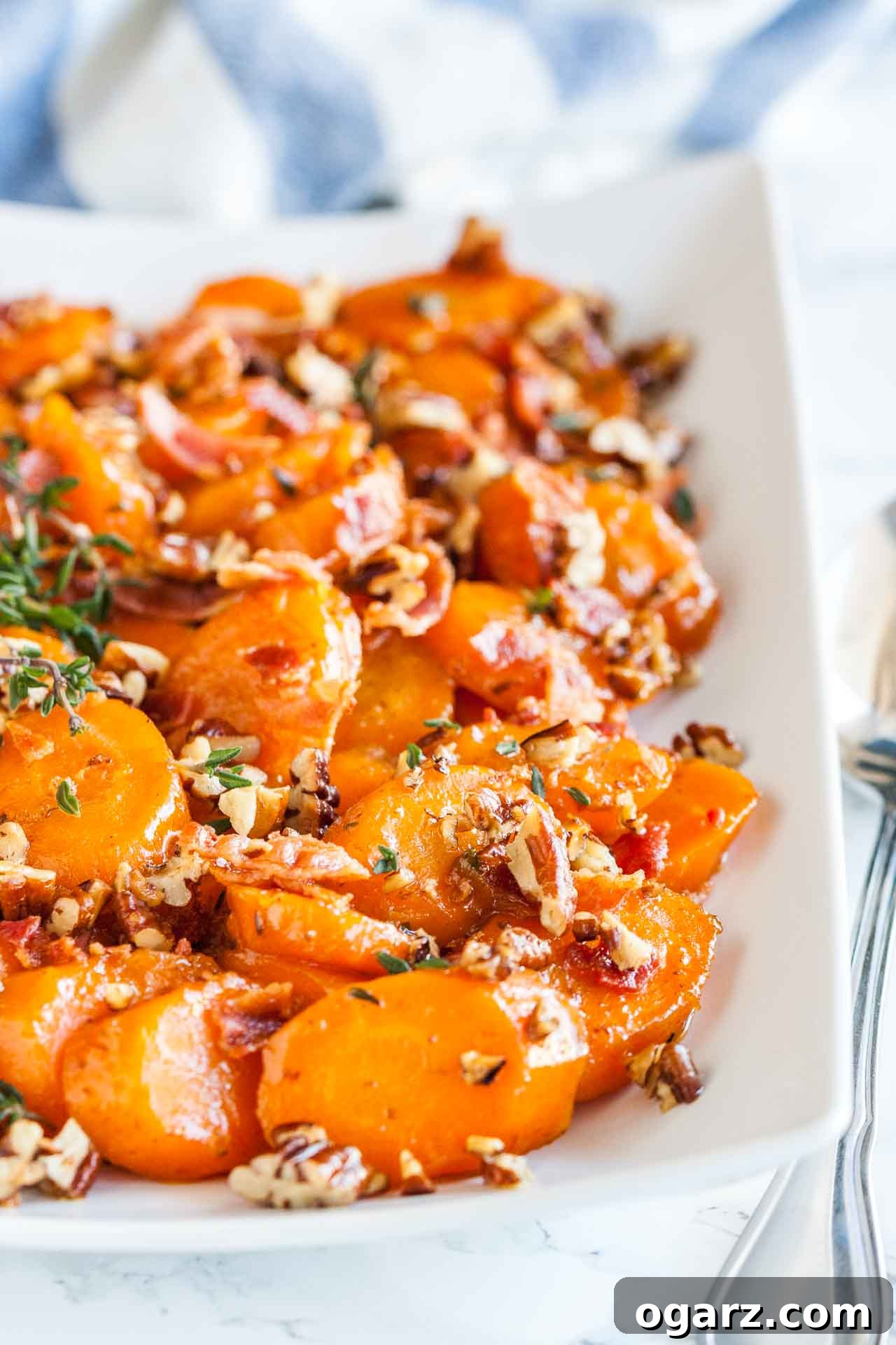 Brown Sugar Carrots: A Sweet Side Dish 2 A rectangular white plate of brown sugar-glazed carrots with bacon and pecans garnished with thyme on a marble surface with a spoon next to it.