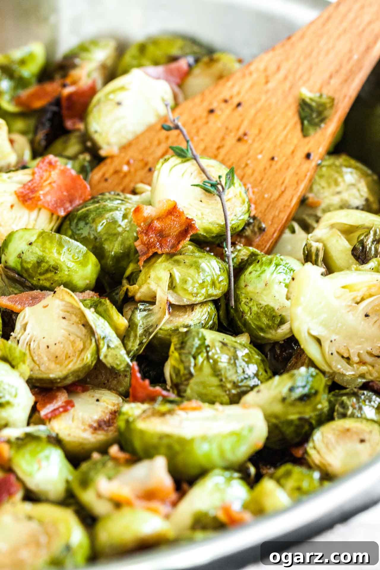 Crispy Brussels Sprouts with Smoked Pork Belly 5 Close-up of roasted brussels sprouts with bacon and a sprig of thyme with a wooden cooking spatula in it.