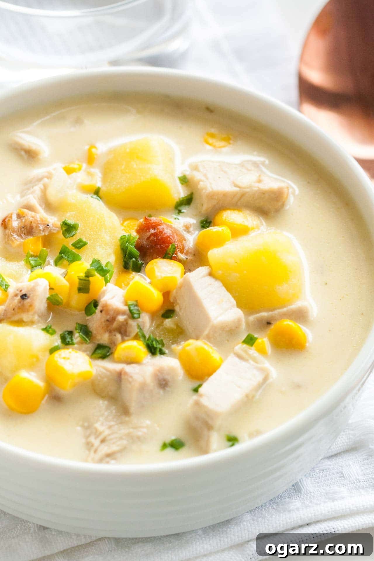 Close-up of a white bowl of creamy leftover turkey soup, garnished with chives on a white dishtowel with a bronze spoon next to it.