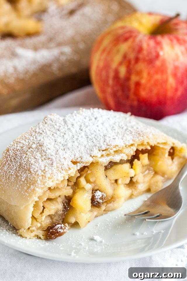 Close-up of a piece of apple strudel topped with powdered sugar on a white plate with a fork on a white dishcloth, with an apple in the background.