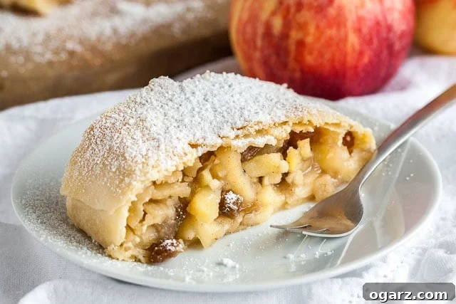 A piece of apple strudel topped with powdered sugar on a white plate with a fork on a white dishcloth, with an apple in the background.