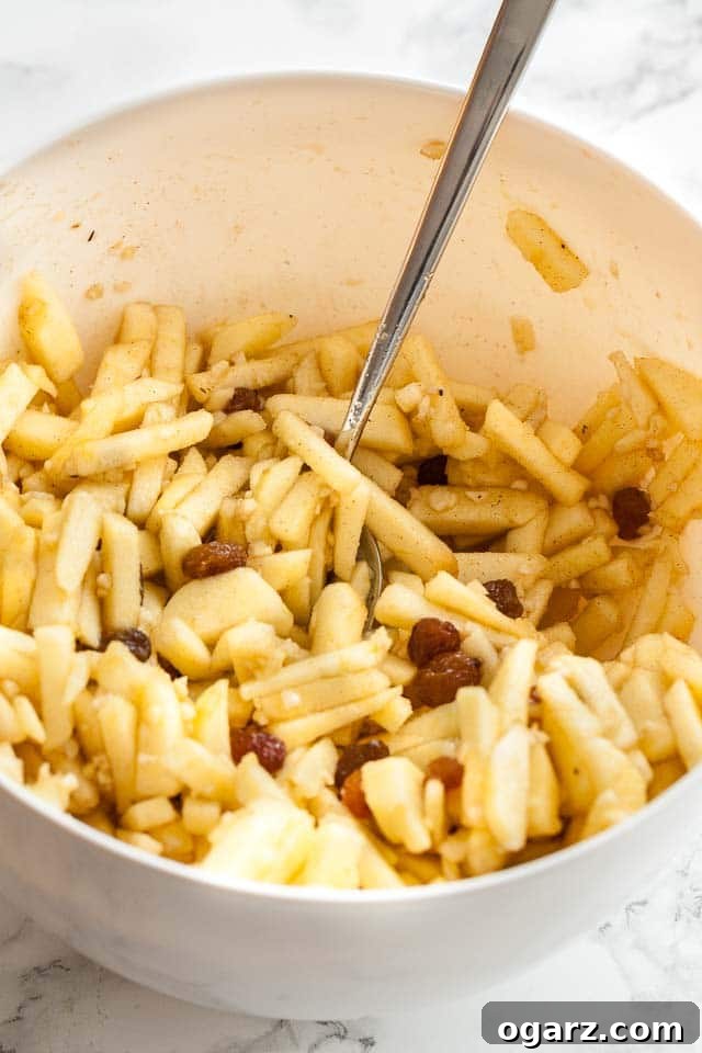 A white bowl containing apples cut into sticks, with raisins and a spoon in it.