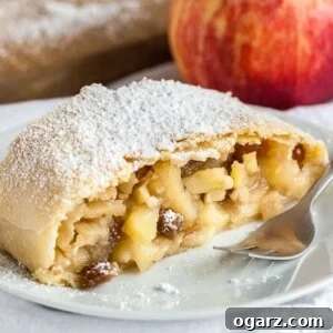 A piece of apple strudel topped with powdered sugar on a white plate with a fork on a white dishcloth, with an apple in the background.