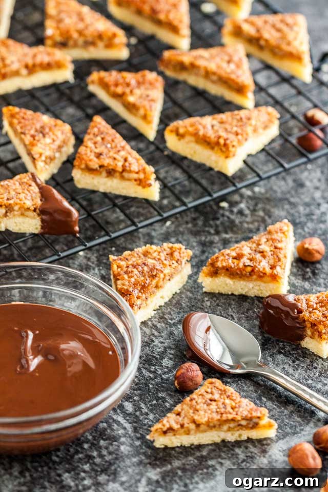 A collection of freshly baked nut corners arranged on and around a black cooling rack, with a glass bowl of melted chocolate ready for dipping.