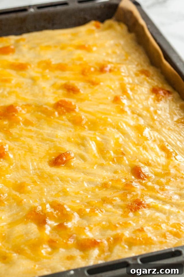 A baking sheet lined with parchment paper, featuring a neatly rolled out shortbread dough base topped with an even layer of vibrant apricot jam.