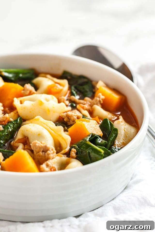 A white bowl of spinach tortellini soup with a wooden spoon next to it on a white tablecloth.