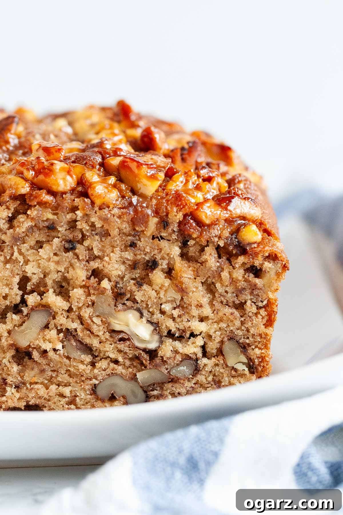 The ultimate moist and delicious banana nut bread, featuring a delectable caramelized nut topping and loaded with walnuts and ripe mashed bananas. A family favorite that's perfect for breakfast or a snack. The best way to use overripe bananas! #bananabread #banananutbread #bananarecipes #bananas #bread #breakfastrecipes Close-up of a loaf of banana nut bread with a slice cut off on a rectangular white plate next to a white and blue dish towel, showing its moist texture and nut topping.