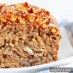 Close-up of a loaf of banana nut bread with a slice cut off on a rectangular white plate next to a white and blue dish towel.