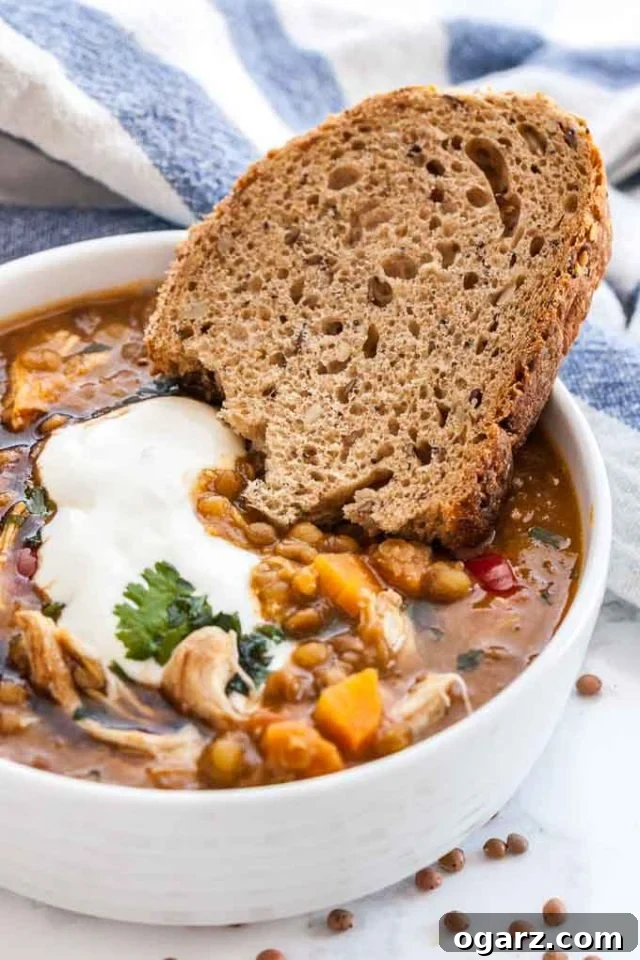 A white bowl of Lentil soup with vegetables and chicken garnished with fresh parsley and a dollop of sour cream, served with a slice of bread. A white and blue dish towel is visible in the background.