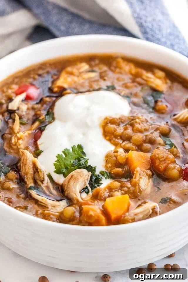 A white bowl of Lentil soup with vegetables and chicken, beautifully garnished with fresh parsley and a swirl of sour cream, resting on a rustic wooden surface with a white and blue dish towel in the background.