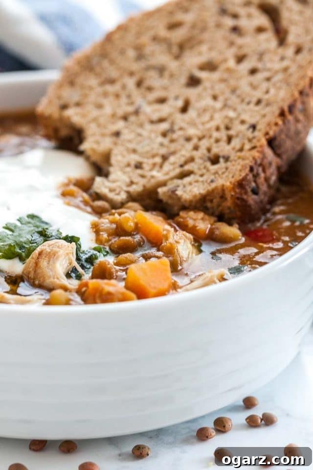 A close-up of our delicious Instant Pot Lentil Soup, ready to be enjoyed with its flavorful vegetables, brown lentils, and shredded chicken. Close-up view of a white bowl filled with Instant Pot Lentil Soup, featuring visible vegetables and shredded chicken, garnished with fresh parsley and sour cream, accompanied by a slice of crusty bread.