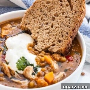 A white bowl of Lentil soup with vegetables and chicken garnished with parsley and sour cream with a slice of bread. A white and blue dish towel is visible.