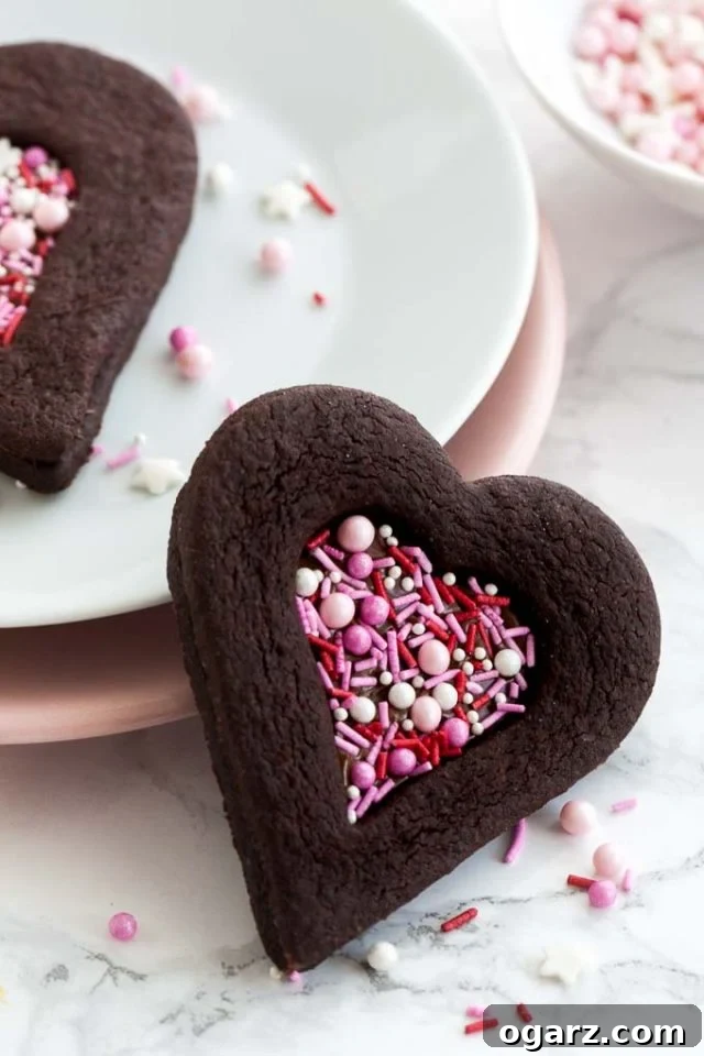 Decadent Chocolate Sugar Cookies 2 Close-up of a heart-shaped chocolate cookie with a heart-shaped indentation filled with pink sprinkles leaning against a stack of plates garnished with sprinkles. There's another cookie on the stack of plates and a small white bowl of pink sprinkles.