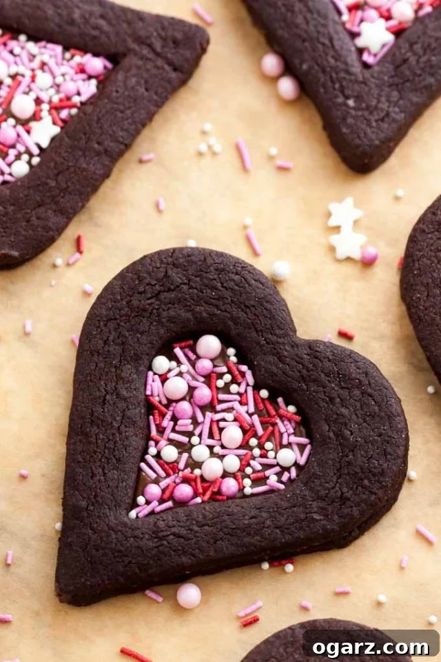 Decadent Chocolate Sugar Cookies 3 Top-down shot of heart-shaped chocolate cookies with a heart-shaped indentation filled with pink sprinkles on parchment paper with some sprinkles.