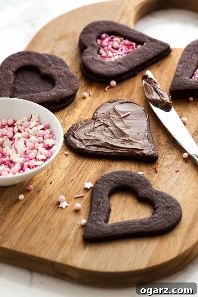 Decadent Chocolate Sugar Cookies 4 A wooden cutting board with heart shaped chocolate cookies in various stages of completion. In the foreground there are the top and bottom part of the cookie and a knife has applied nutella to the bottom. The top has a smaller heart cut out. There's a small bowl of sprinkles next to it.