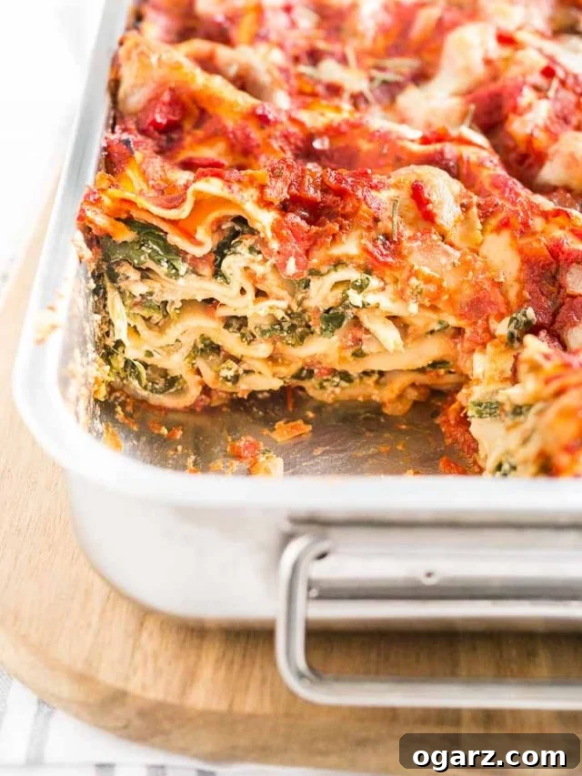 Delicious Spinach Lasagna - Perfect Vegetarian Comfort Food A metal baking pan with spinach lasagna on a wooden cutting board. There's a piece of it missing, showcasing the layers.