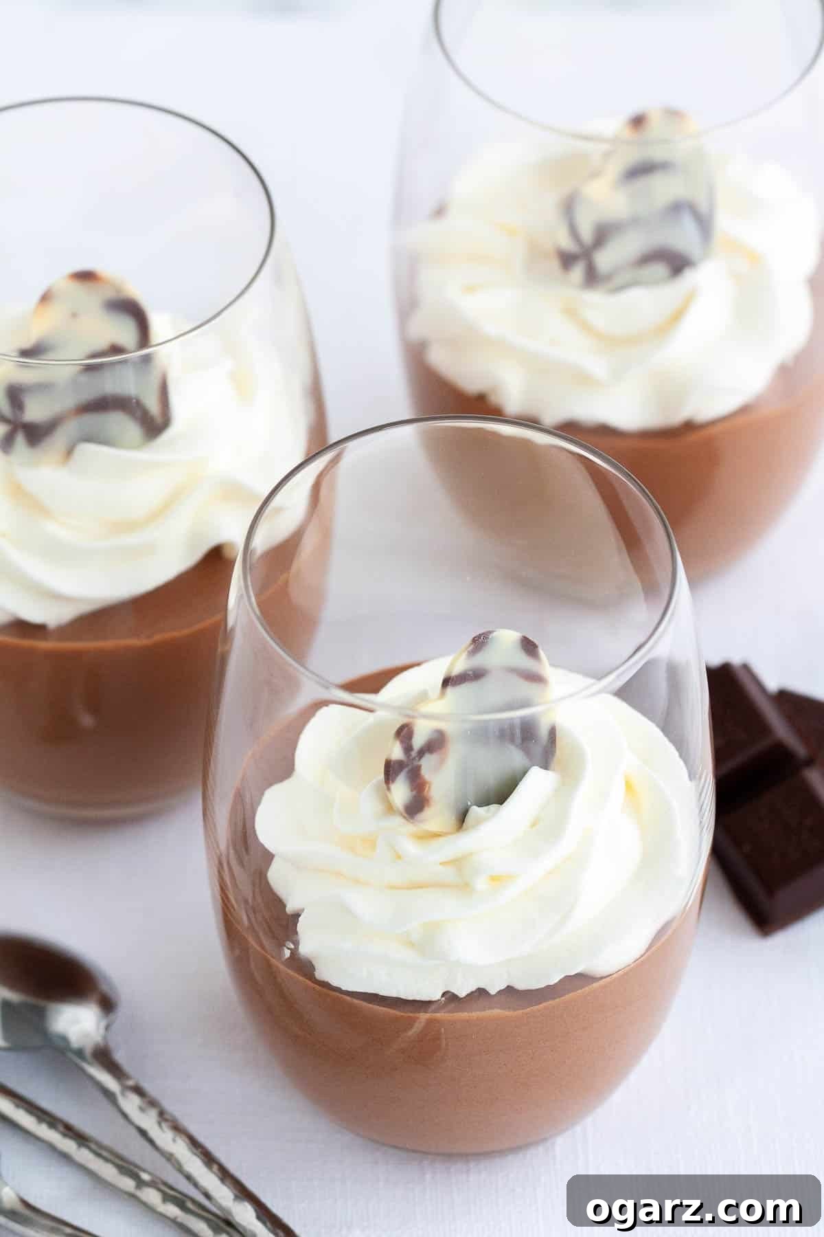 This easy Chocolate Mousse recipe is super simple to make from scratch and deliciously light and fluffy. An eggless mousse recipe that is so airy and chocolatey and can be enjoyed by everyone. It's fancy enough for a party but easy enough to make whenever you have a craving for chocolate! Three glasses of chocolate mousse, topped with whipped cream next to pieces of chocolate and a spoon.