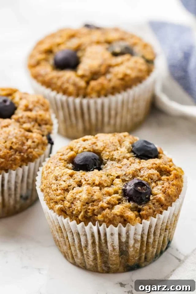 Golden Banana Blueberry Muffins 2 Three golden-brown banana blueberry muffins arranged on a white marble surface, with a charming white and blue dishtowel gently placed beside them. The muffins showcase their characteristic domed tops and visible blueberries.