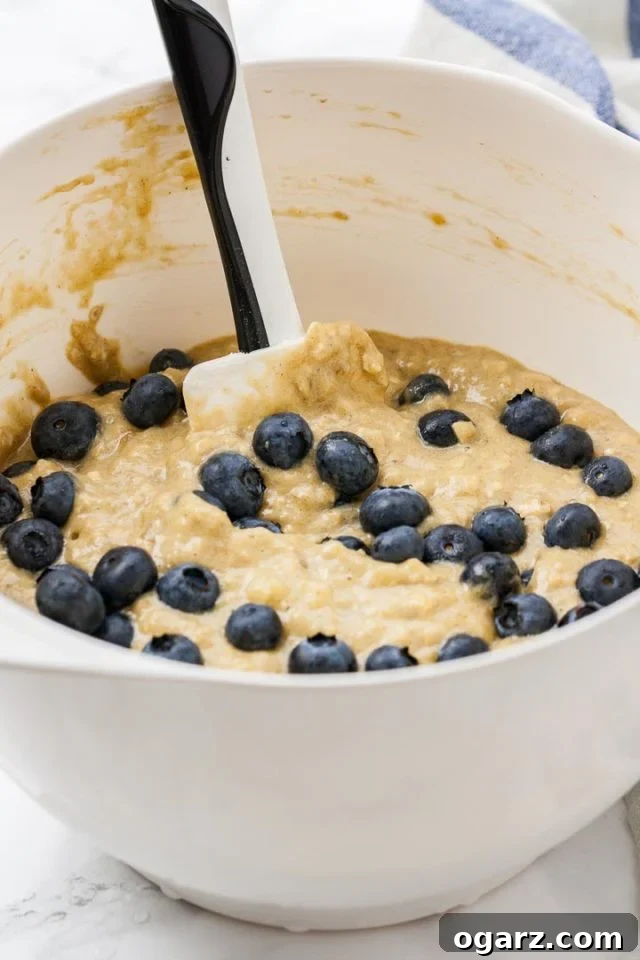Golden Banana Blueberry Muffins 3 A large white mixing bowl filled with a golden muffin batter, generously mixed with fresh blueberries. A black and white spatula rests in the bowl.