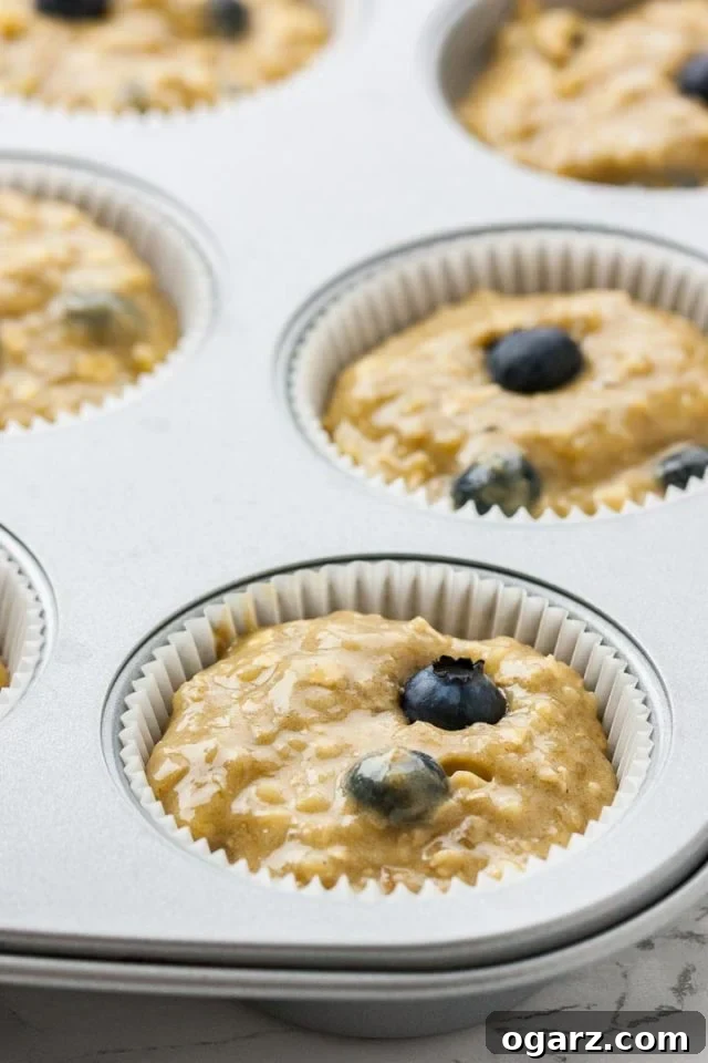 Golden Banana Blueberry Muffins 4 A 12-cup muffin pan lined with white paper baking cups, each filled with a dollop of muffin batter mixed with vibrant blueberries, ready for baking.