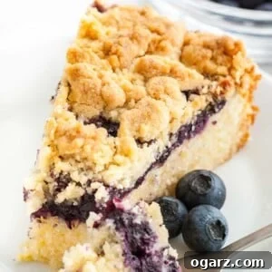 A slice of blueberry breakfast cake with streusel topping on a white plate, garnished with fresh blueberries, with a fork beside it.