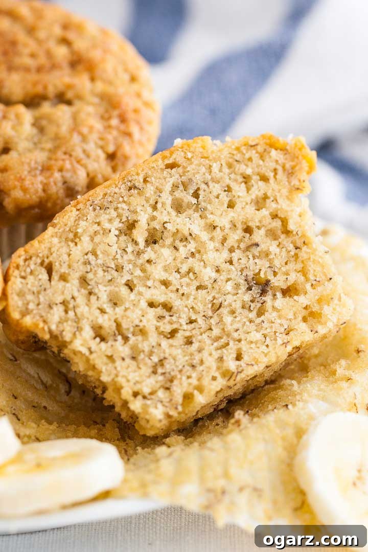 Foolproof Banana Muffins 3 A banana muffin unwrapped and halved, showcasing its moist, fluffy interior. Another muffin and a blue and white striped towel are visible in the soft focus background.