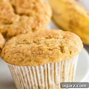 Close-up of a muffin on a white plate with several muffins and a banana in the background.