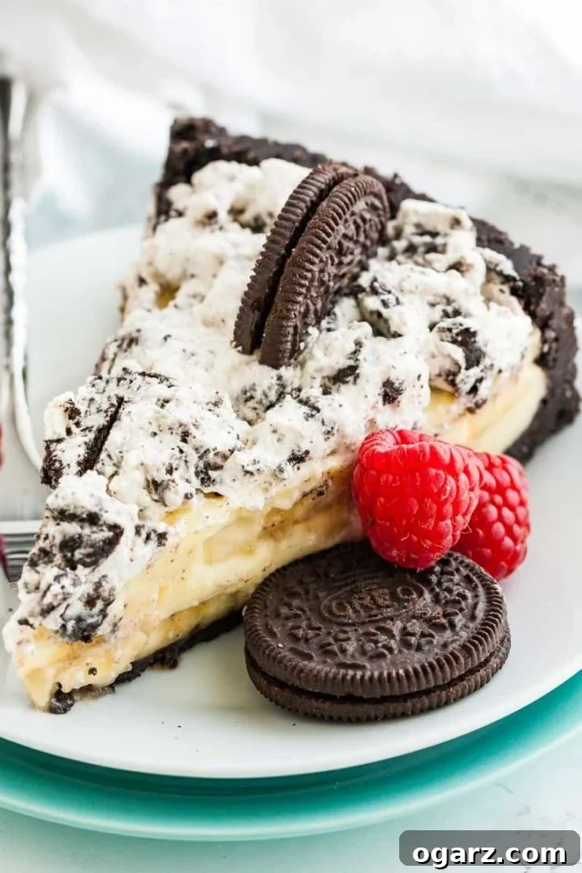 A slice of Oreo banana cream cake on a white plate with a fork, garnished with an oreo cookie and two raspberries.