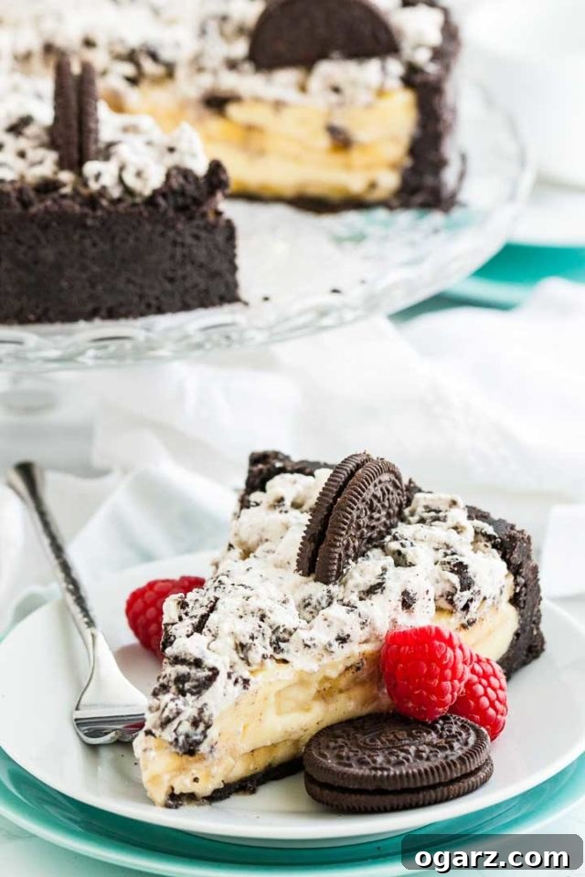 A slice of oreo banana cream cake on a plate with a fork, garnished with raspberries and an oreo cookie. In the background, there\'s a glass serving dish with the rest of the cake on it.